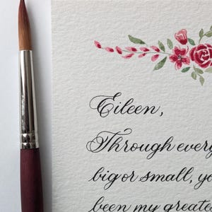 May include: A watercolor painting of a floral design with red roses and green leaves, and calligraphy text. A paintbrush with a maroon handle and silver ferrule is on the left. The text reads: "Eileen, Through every big or small, you been my greatest Forty ruby years."