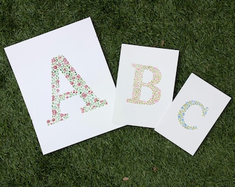 May include: Three white art prints featuring the letters A, B, and C. The letters are decorated with floral patterns in shades of red, pink, green, and blue. The prints are arranged on a bed of green grass.