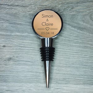 Pode incluir: Um elegante stopper de garrafa de vinho personalizado com acabamento prateado. Apresenta um topo redondo bege com os nomes "Simon & Claire", um símbolo de coração e a data "05.04.18" em texto preto. Com vedação de borracha preta.