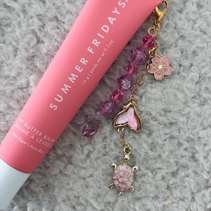 May include: Pink lip balm tube with a gold chain and charms, including a pink flower, a butterfly, and a turtle. The tube is labelled "Summer Fridays" and "Lip Butter Balm".