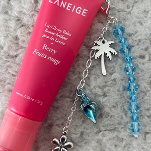 May include: A pink tube of LANEIGE Lip Glowy Balm in Berry Fruits rouge flavour with a silver charm attached. The charm has a blue seashell, a silver flower, a silver palm tree, and a strand of blue crystal beads.