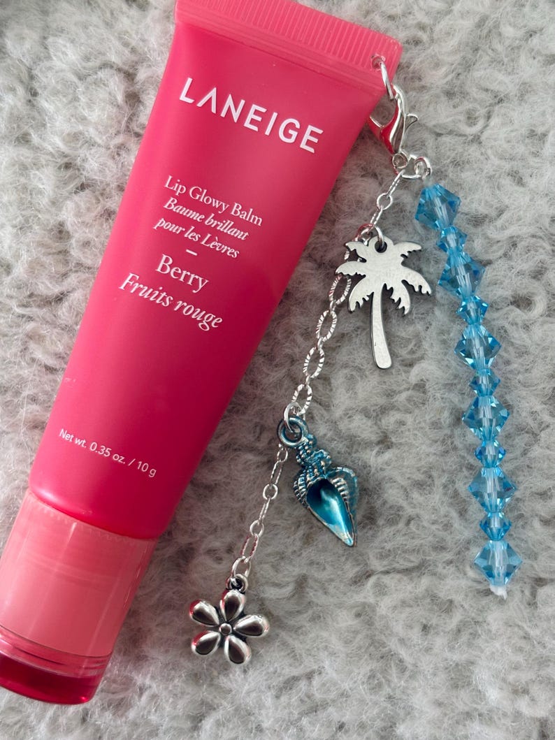 May include: A pink tube of LANEIGE Lip Glowy Balm in Berry Fruits rouge flavour with a silver charm attached. The charm has a blue seashell, a silver flower, a silver palm tree, and a strand of blue crystal beads.