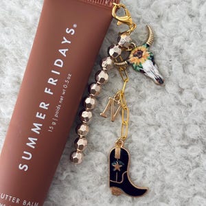 May include: A brown tube of lip balm with the text "SUMMER FRIDAYS." and "15 g | poids net wt 0.5 oz". The tube is attached to a gold charm bracelet with a cowboy boot, a skull with a sunflower, and a gold letter "M".