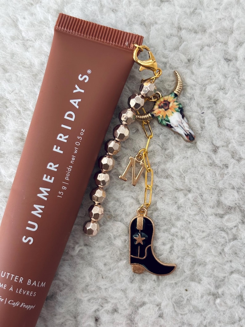 May include: A brown tube of lip balm with the text "SUMMER FRIDAYS." and "15 g | poids net wt 0.5 oz". The tube is attached to a gold charm bracelet with a cowboy boot, a skull with a sunflower, and a gold letter "M".