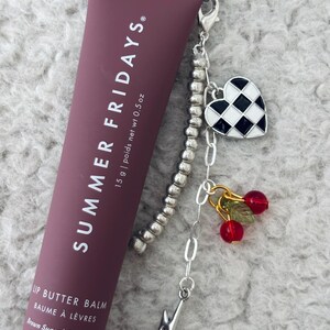 May include: A brown tube of lip balm with the text "SUMMER FRIDAYS" and "LIP BUTTER BALM BAUME &Agrave; L&Egrave;VRES Brown Sugar | Sucre Brun" on the front. The tube is attached to a silver charm bracelet with a black and white checkered heart charm, a lightning bolt charm, and two red cherry charms.