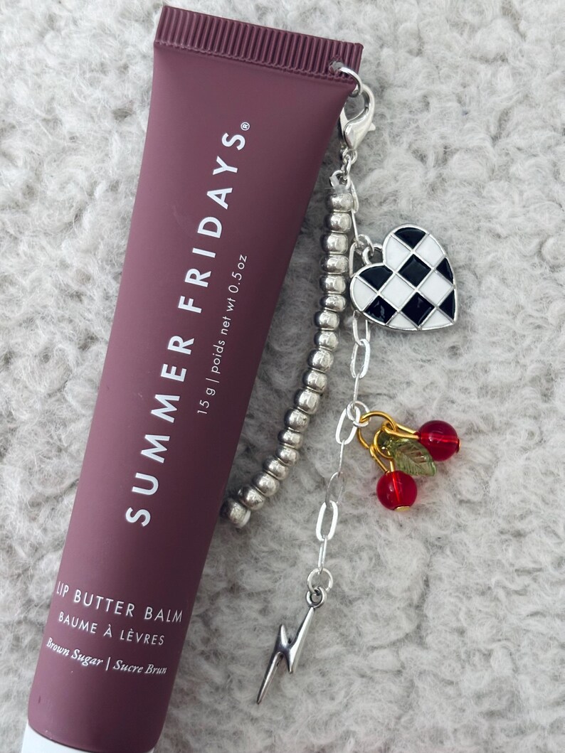 May include: A brown tube of lip balm with the text "SUMMER FRIDAYS" and "LIP BUTTER BALM BAUME &Agrave; L&Egrave;VRES Brown Sugar | Sucre Brun" on the front. The tube is attached to a silver charm bracelet with a black and white checkered heart charm, a lightning bolt charm, and two red cherry charms.