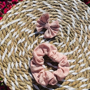 Puede incluir: Dos scrunchies para el pelo con estampado rosa y blanco, uno es un lazo y el otro es un scrunchie redondo. Los scrunchies están sobre una estera de paja tejida.