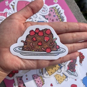 May include: A white sticker with a glittery finish depicting a brown cake with red frosting and gummy worms on a white plate.