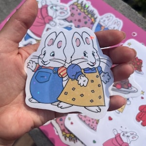 May include: Two white rabbit characters with glitter accents. The rabbit on the left is wearing blue overalls and a red and white striped shirt. The rabbit on the right is wearing a blue shirt and a yellow dress with white polka dots.