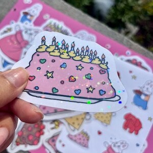 Peut inclure: Un autocollant de g&acirc;teau rose et blanc avec des paillettes, des bougies jaunes et des vermicelles color&eacute;s. L'autocollant est sur un fond rose avec d'autres autocollants.