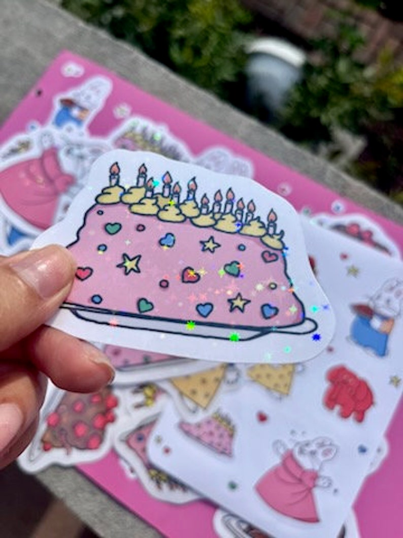 Peut inclure: Un autocollant de g&acirc;teau rose et blanc avec des paillettes, des bougies jaunes et des vermicelles color&eacute;s. L'autocollant est sur un fond rose avec d'autres autocollants.