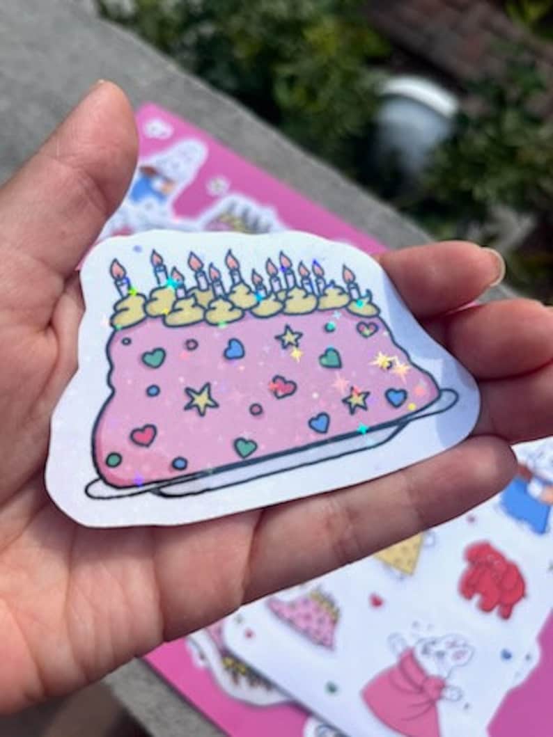 Peut inclure: Un autocollant de g&acirc;teau d'anniversaire rose et blanc avec des bougies color&eacute;es, des p&eacute;pites et des c&oelig;urs. Le g&acirc;teau est d&eacute;cor&eacute; de paillettes et a une finition holographique.