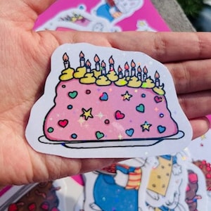 May include: A pink and white sticker of a birthday cake with yellow frosting, candles, and sprinkles. The cake is decorated with hearts and stars.