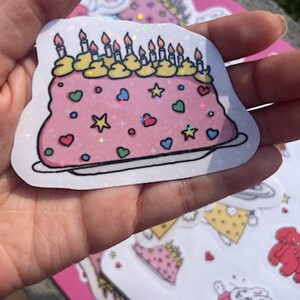 Peut inclure: Un g&acirc;teau rose et blanc avec un gla&ccedil;age jaune et des bougies. Le g&acirc;teau est d&eacute;cor&eacute; de c&oelig;urs, d'&eacute;toiles et de p&eacute;pites. Le g&acirc;teau est sur une assiette blanche. Le g&acirc;teau est un autocollant avec une finition scintillante.