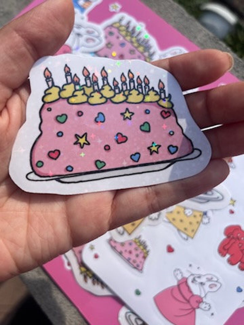 Peut inclure: Un g&acirc;teau rose et blanc avec un gla&ccedil;age jaune et des bougies. Le g&acirc;teau est d&eacute;cor&eacute; de c&oelig;urs, d'&eacute;toiles et de p&eacute;pites. Le g&acirc;teau est sur une assiette blanche. Le g&acirc;teau est un autocollant avec une finition scintillante.