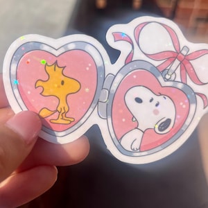 May include: Two pink heart-shaped locket stickers with glitter accents. One features a cartoon image of Snoopy, the other features Woodstock.