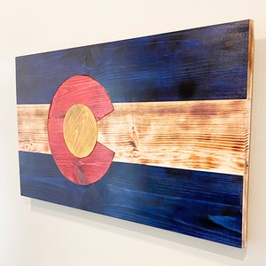 May include: A wooden wall art piece depicting the Colorado state flag. The flag is painted in shades of blue, red, and yellow on a wooden surface. The flag is mounted on a wall.
