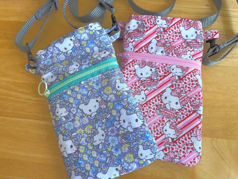 May include: Two small crossbody bags with adjustable gray straps. One bag is patterned with Hello Kitty characters and flowers on a blue background, and the other has a pink and red Hello Kitty design. Both have zippered closures.
