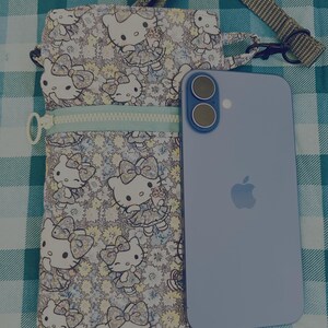 May include: A floral print crossbody bag featuring a Hello Kitty design, with a light blue zipper and strap. A light blue smartphone with a dual-camera setup is next to the bag, resting on a checkered surface.