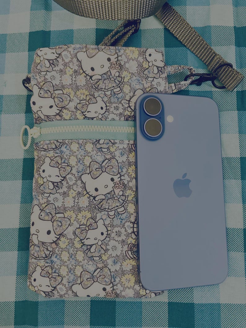 May include: A floral print crossbody bag featuring a Hello Kitty design, with a light blue zipper and strap. A light blue smartphone with a dual-camera setup is next to the bag, resting on a checkered surface.