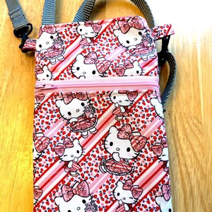 May include: A rectangular crossbody bag featuring a repeating pattern of a white cat with a pink bow and dress, set against a background of red flowers and pink stripes. The bag has a pink zipper, a gray adjustable strap, and a gray handle.