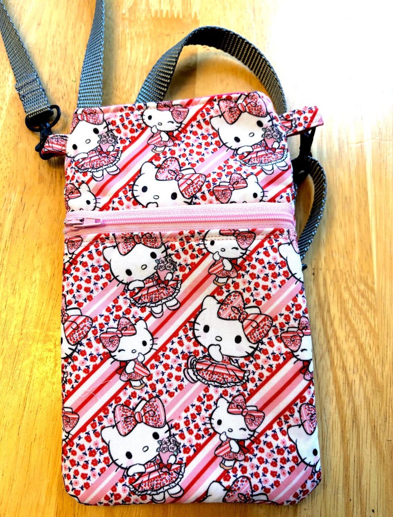 May include: A rectangular crossbody bag featuring a repeating pattern of a white cat with a pink bow and dress, set against a background of red flowers and pink stripes. The bag has a pink zipper, a gray adjustable strap, and a gray handle.