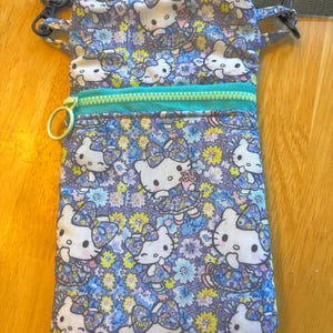May include: A rectangular crossbody bag with a floral and Hello Kitty print. The bag features a teal zipper, a gold-colored zipper pull, and a gray adjustable strap. The pattern includes white Hello Kitty figures, blue, purple, and yellow flowers.