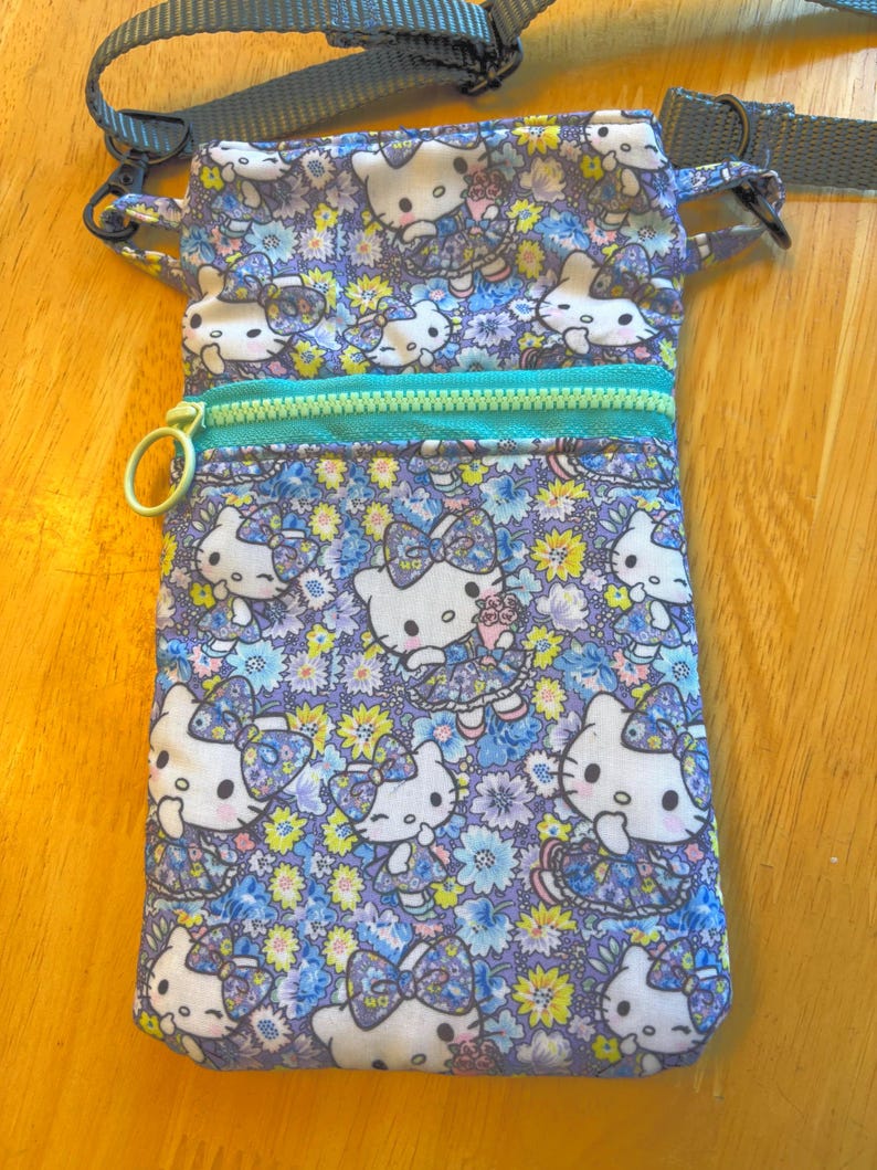 May include: A rectangular crossbody bag with a floral and Hello Kitty print. The bag features a teal zipper, a gold-colored zipper pull, and a gray adjustable strap. The pattern includes white Hello Kitty figures, blue, purple, and yellow flowers.