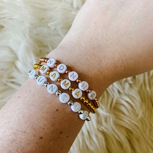 May include: Three beaded bracelets spelling out "AUNTIE", "MAMA", and "NONNA" in white letter beads. The bracelets are on a wrist, with gold, silver, and copper-coloured beads. The background is a fluffy, white surface.