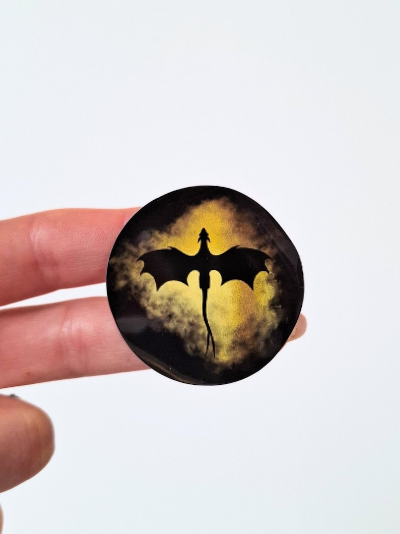 May include: A circular pin featuring a black silhouette of a dragon with outstretched wings centered against a vibrant yellow and gold cloud-like background. The design is encased in a smooth, glossy finish that reflects light across the surface.