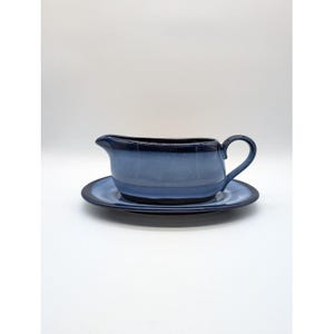 May include: A blue ceramic gravy boat with a matching saucer. The gravy boat has a handle and a spout. The set is a gradient of blue hues, with a darker blue rim. The set is on a white background.