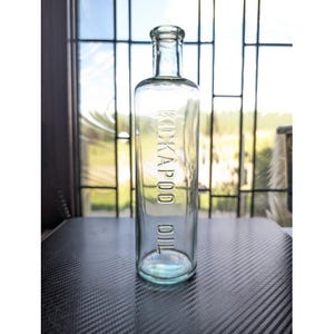 May include: Clear glass bottle with the embossed text "KICKAPOO OIL". The bottle has a rectangular shape with rounded edges and a narrow neck. The bottle is displayed in front of a window.
