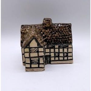 May include: A ceramic miniature house figurine featuring a cream-colored base with dark brown painted timber framing and a textured, dark brown glazed roof. The structure includes a central chimney and small windows with a cross-hatch pattern.