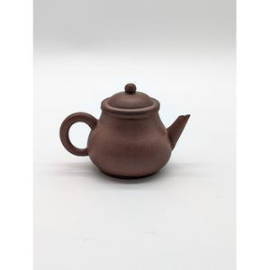 May include: A brown ceramic teapot with a rounded body, a curved handle, and a spout. The lid has a small knob on top. The teapot is designed for brewing tea and has a classic, simple design.