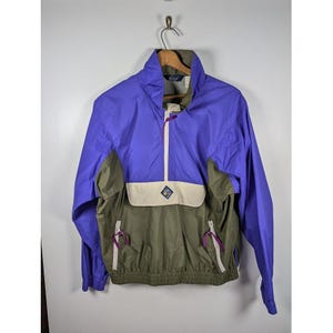 May include: A vintage anorak-style jacket in purple, olive green, and cream. The jacket features a half-zip closure, a front pocket with a diamond-shaped logo, and purple zipper pulls. The jacket is hanging on a metal hanger.
