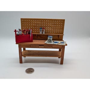 May include: Miniature wooden workbench with a red toolbox, tools, and paint cans. The workbench has a pegboard back, a drawer, and a lower shelf. A coin is in the foreground.