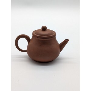 May include: A small, round, brown teapot with a matte, textured surface. The teapot features a curved handle on the left side, a short, pointed spout on the right, and a circular lid with a small, rounded knob handle centered on the top.