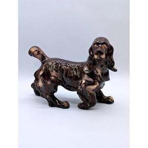 May include: A ceramic dog figurine in shades of brown and bronze. The dog is posed in a standing position with its tail up and ears hanging down. The figurine has a glossy finish and is likely a decorative item.
