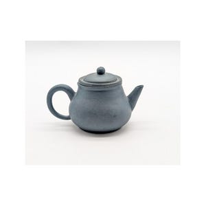 May include: A small, blue-grey ceramic teapot with a rounded body, a curved handle, and a spout. The lid has a small knob on top. The teapot is likely used for brewing tea and has a matte finish.