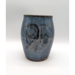 May include: A cylindrical ceramic mug with a speckled blue and grey glaze. The exterior features a dark, abstract brushstroke design in black and charcoal grey that wraps around the center of the vessel. The rim is finished with a dark brown edge.