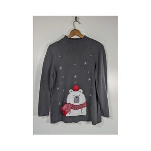 May include: A charcoal gray long-sleeve sweater with a mock neck collar. The front features a white bear graphic wearing a red and white patterned scarf, a small red bird on its head, and embroidered white stars and dots resembling falling snow.
