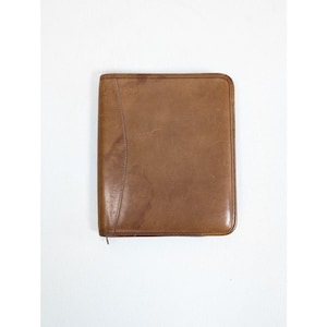 May include: A rectangular brown leather portfolio case with a smooth, worn texture and rounded corners. The case features a vertical stitched seam detail along the left side and a zipper closure that runs along the perimeter of the document holder.