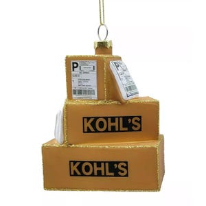 May include: A decorative Christmas ornament shaped like a stack of three cardboard boxes. The boxes are gold with black text that reads "KOHL'S". Each box has a shipping label. The ornament is trimmed with gold glitter.