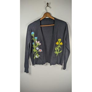 May include: A dark gray cardigan sweater with embroidered floral designs in shades of blue, pink, yellow, and white. The open-front sweater is hanging on a wooden hanger against a light gray wall.