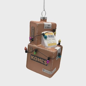 May include: A Christmas ornament shaped like two stacked cardboard boxes with the Kohl's logo. The boxes are brown with glitter and are decorated with a string of colorful Christmas lights and a mail label.