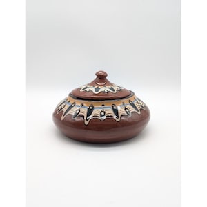 May include: A brown ceramic lidded bowl with a decorative pattern in white, black, and blue. The bowl has a rounded shape with a matching lid and a small knob. The design features a repeating wave-like pattern around the bowl's rim.