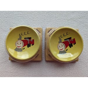 May include: Two yellow ceramic plates with a cartoon train design. The train is red and black with a smiling face. The plates are packaged on cardboard backing.