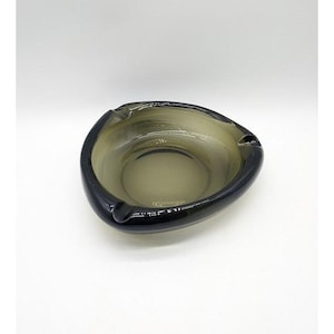 May include: A smoke-colored glass ashtray with a rounded triangular shape and three distinct notches along the thick, dark-tinted rim. The center of the bowl is translucent, transitioning to a deeper, darker tone toward the outer edges.