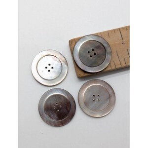 May include: Four large, round, iridescent buttons with four holes each. The buttons are a pearlescent color, with a subtle sheen. One button is partially on a wooden ruler.