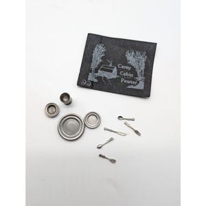 May include: Miniature pewter tableware set, including plates, cups, spoons, a fork, and a knife. A dark gray square with the text "Carey Cabin Pewter" and a cabin illustration is also included. The set is arranged on a white surface.